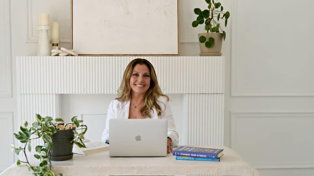 Woman Angela Masuzzo working on a laptop at a white desk with plants and books
